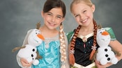 Two young girls dressed as Anna and Elsa hold plush Olaf toys and pose for a studio portrait against a standard backdrop