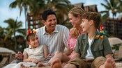 A dad, a mom and their 2 young daughters relaxing on a beach area in front of Aulani, A Disney Resort and Spa