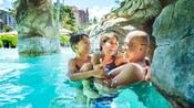 A smiling couple holds each other and their young son in a pool at Aulani Resort