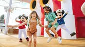 Two children smiling and leaping with Captain Minnie Mouse and Captain Mickey Mouse on an upper deck of a Disney Cruise Line ship