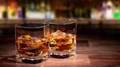 Two glasses of whiskey with ice sit on a wooden surface.