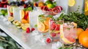 A variety of beverages arranged on a marble countertop, garnished with fresh fruits, herbs and ice.