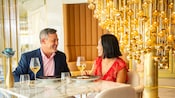 A couple enjoying an upscale meal in front of a chandelier of cascading balls in an elegant restaurant aboard a Disney cruise ship