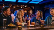 Huéspedes sonriendo mientras disfrutan de comida y bebidas en el bar de Periscope Pub en el Disney Treasure