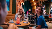 Hóspedes saboreando bebidas em uma mesa enquanto músicos tocam no palco atrás deles no Skipper Society, um lounge no Disney Treasure