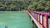 A long wooden jetty extends over the water toward a sandy cove, framed by dense jungle hillsides