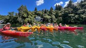 Group of Guests in kayaks floating on calm water near a forested shoreline waving for the camera