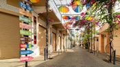 Calle peatonal en Puerto Plata bordeada por sombrillas sobre la cabeza, florecientes viñedos y letreros comerciales españoles
