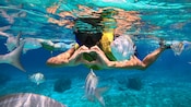 A woman snorkeling and making a heart shape with her hands, surrounded by several fish