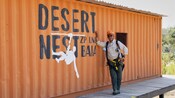 A smiling man in a zip line harness leans against a structure on which are the words ‘Desert Nest Zip Line Baja’