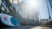 Uma prancha de stand-up paddle e um remo encostados em uma grade de metal em uma passarela próxima a um porto