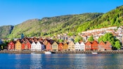 Colorful wooden buildings lined along a waterfront with forested mountains in the background