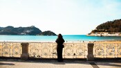 A woman standing at an ornate railing overlooking a beach with hills and water