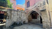 An ancient stone fountain with spouts dripping water next to an arched entryway surrounded by buildings