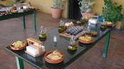 A long table set with 2 napkin holders and terracotta bowls holding sliced baguette, olives, chips and a tray of small sample cups