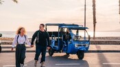 A couple walking near a Tuk Tuk motorized vehicle as the sun sets on the Málaga coastal