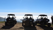 A trio of off road buggies parked on a ridge looking at mountains surrounded by a low layer of clouds