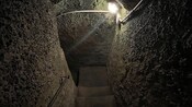 Narrow stone staircase in an underground passage with rough walls and an overhead light