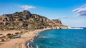 A sandy beach with sun loungers stretching along the coastline, bordered by calm waters and rocky hills