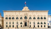 The ornate facade of the Auberge de Castille in Valletta, Malata reveals intricate columns and a flag flying from the rooftop