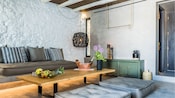 A living room with a couch, floor cushions, a lamp and a coffee table with 2 bottles of wine and fruit