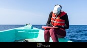 Un Visitante con un chaleco salvavidas escribiendo en un portapapeles a bordo de un pequeño bote en aguas abiertas con las palabras “National Geographic Day Tours”