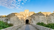 A historic stone fortress with a central arched entrance, flanked by 2 round towers and connected by a stone bridge