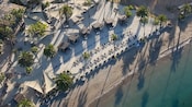 An aerial view of a beach resort featuring several cabanas with wooden roofs, lounge chairs, umbrellas and palm trees on the sand