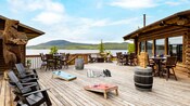 Una plataforma junto al lago entre dos cabañas de madera con mesas, sillas, juegos al aire libre y una fogata