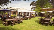 Outdoor dining area with wooden tables and chairs on a grassy lawn with umbrellas and trees