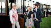 A couple engaging with a conductor figure wearing a vest and a hat outside the vintage road train