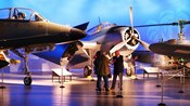A man and 2 kids talking to a guide near several aircraft on display at the Air Force Museum of New Zealand