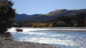 A jet boat speeding down a river surrounded by trees, rocky shores and rolling hills