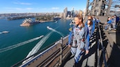 Visitantes caminando por el Puente del Puerto de Sídney con la Ópera de Sídney detrás de ellos