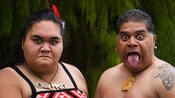 Two Māori people wearing traditional clothing and jewelry and demonstrating pūkana, which are bold facial expressions