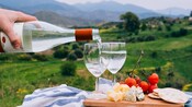 A hand pouring wine into 2 stemmed glasses set next to a serving board with cheese, cherry tomatoes and crackers