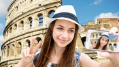 A young woman holding her smart phone and posing for a selfie in front of the Roman Colosseum 