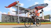 A man riding an electric bicycle next to a jet plane that is painted like a shark