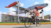 A man riding an electric bicycle next to a jet plane that is painted like a shark