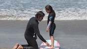 Una niña parada en una tabla de surf cerca de la costa recibiendo instrucciones sobre cómo surfear