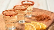 Three shot glasses of tequila with tajin on the rim beside 3 slices of orange.