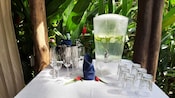 A table in a cabana with a champagne bucket, a cucumber infused water dispenser, champagne glasses and water glasses