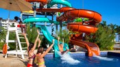 2 kids cheering as a man splashes down a waterslide at an outdoor pool