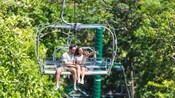 A couple pointing at their surroundings while riding a chair lift amid trees