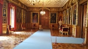 Una sala con paneles de madera que exhibe pinturas de retratos, con pisos de madera, candelabros que cuelgan de un techo tallado y el busto de mármol de un hombre