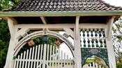 A gate with a tile roofed overhang and an arch above its picket fence that features animal carvings and has German words written on it