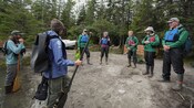 Grupo de personas con chalecos salvavidas y equipo para el aire libre reunidos en un área boscosa, preparando una actividad de canotaje