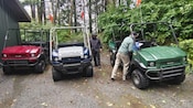 Three open air jeeps parked at the edge of a forest, with a man retrieving a notebook from one of them and another man standing nearby