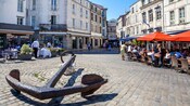 A European square with cobblestone, cafes and a large metal anchor at the center