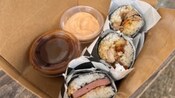 Una caja de un restaurante con comida para llevar que incluye tres&nbsp;rollos de sushi Spam y dos&nbsp;salsas para untar